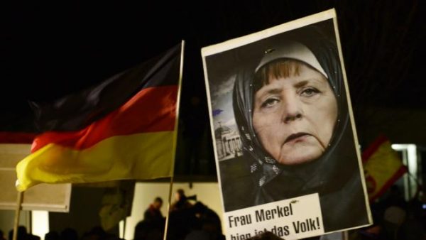 A protestor holds a poster with an image of German Chancellor Angela Merkel wearing a headscarf in front of the Reichstag building with a crescent on top during a rally of the group Patriotic Europeans against the Islamization of the West, or PEGIDA, in Dresden, Germany, Monday, Jan. 12, 2015. (AP Photo/Jens Meyer)
